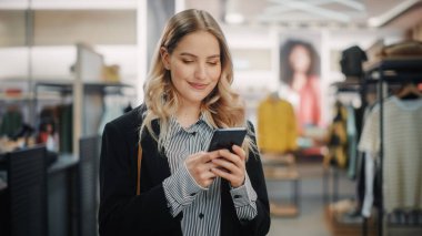 Güzel Gülümseyen Kadın Müşteri Alışverişi, Akıllı Telefon Kullanımı, İnternet Taraması, İnternette Karşılaştırma, Şık Kıyafetler Seçme. Moda Dükkanı, Renkli Markalar, Sürdürülebilir Tasarımlar