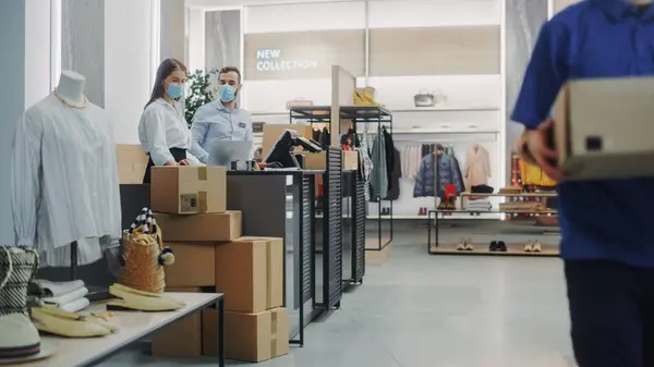 Clothing Store Checkout Cashier Counter: Female and Male Retail Sales Managers wearing Protective Face Masks Give Package to Online Order Delivery Person. Designer Brands Available to Buy on Internet