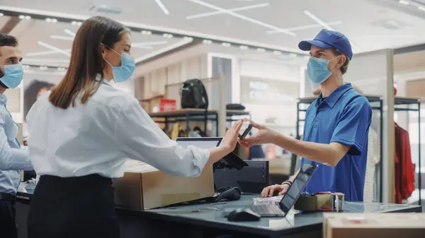 Clothing Store Checkout Counter: Retail Sales Managers wearing Protective Face Masks Give Package to Courier for Online Order Delivery. Trendy Designer Brands Available on Internet.
