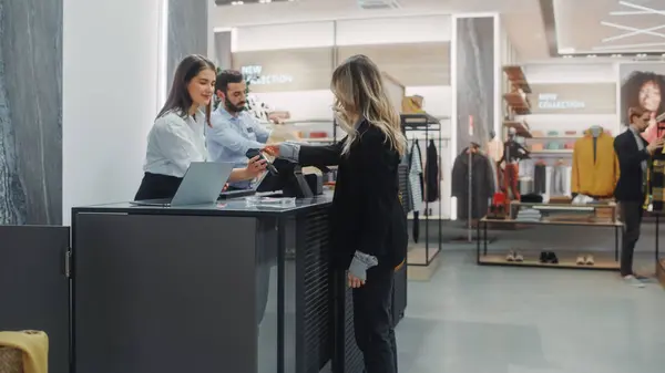 Clothing Store Checkout Cashier Counter: Beautiful Young Woman Buys Blouse from Friendly Retail Sales assistant, Paying with Contactless Credit Card. Stylish Fashion Shop with of Designer Brands