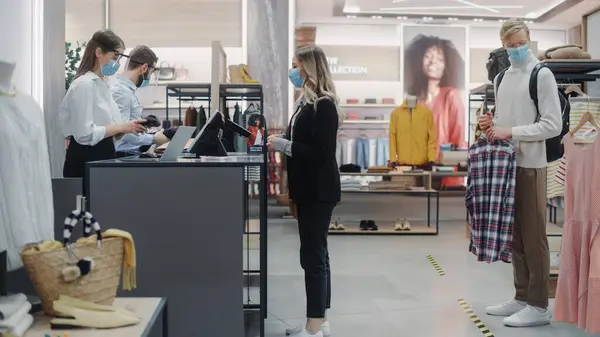 Clothing Store: Young Woman At Counter Buys Clothes from Friendly Retail Sales assistant, Paying with Contactless Credit Card. Everybody Wearing Face Masks. Trendy Fashion Shop with of Designer Brands