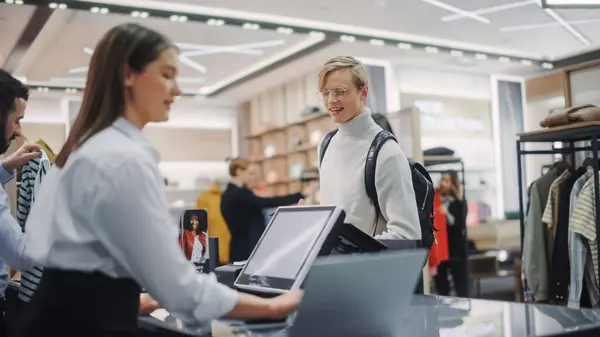 stock image Clothing Store: Young Man At Checkout Counter Buys Clothes Paying with Contactless NFC Smartphone, Friendly Retail Sales Assistan Packs Merchandise. Contemporary Fashion Shop with of Designer Brand