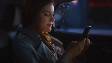 Stylish Female, Gece Bir Taksinin Arka Koltuğu İçinde Eve Dönüş Yapıyor. Smartphone kullanan ve pencereden dışarı bakan güzel bir kadın. Şehir caddesinde neon ışıklarıyla çalışan bir arabada..