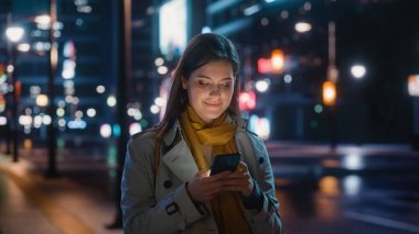 Trençkot giymiş güzel bir kadının portresi modern şehir caddesinde neon ışıklarıyla yürüyor. Akıllı Telefon Kullanan Çekici Kadın ve Şehir Sineması Çevresine Bakış.