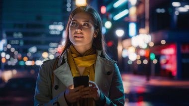 Trençkot giymiş güzel bir kadının portresi modern şehir caddesinde neon ışıklarıyla yürüyor. Akıllı Telefon Kullanan Çekici Kadın ve Şehir Sineması Çevresine Bakış.