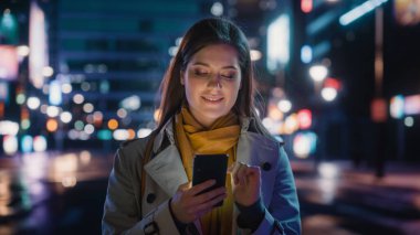 Trençkot giymiş güzel bir kadının portresi modern şehir caddesinde neon ışıklarıyla yürüyor. Akıllı Telefon Kullanan Çekici Kadın ve Şehir Sineması Çevresine Bakış.