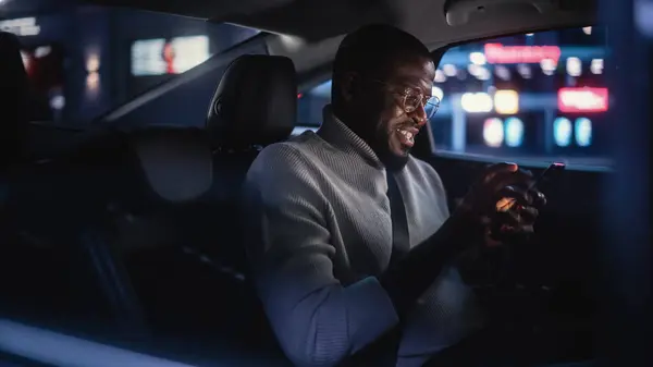ハッピー ブラック グラスが夜のタクシーの後部座席で帰宅 働くネオン サインと都市街の車の中でスマートフォンと笑顔を使用するハンサムな男性 — ストック写真