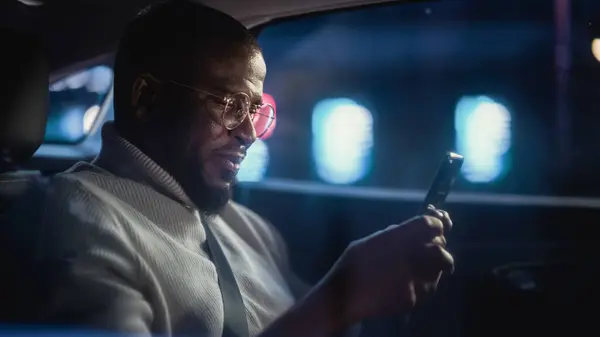 ハッピー ブラック グラスが夜のタクシーの後部座席で帰宅 働くネオン サインと都市街の車の中でスマートフォンと笑顔を使用するハンサムな男性 — ストック写真