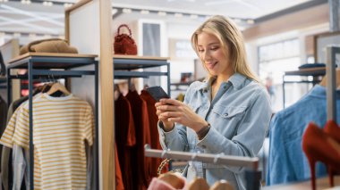 Güzel Gülümseyen Kadın Müşteri Alışverişi, Akıllı Telefon Kullanımı, İnternet Taraması, İnternette Karşılaştırma, Şık Kıyafetler Seçme. Moda Dükkanı, Renkli Markalar, Sürdürülebilir Tasarımlar