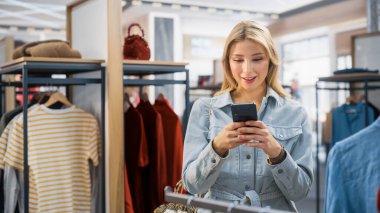 Güzel Gülümseyen Kadın Müşteri Alışverişi, Akıllı Telefon Kullanımı, İnternet Taraması, İnternette Karşılaştırma, Şık Kıyafetler Seçme. Moda Dükkanı, Renkli Markalar, Sürdürülebilir Tasarımlar