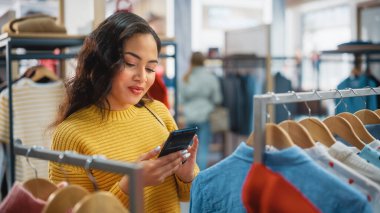 Güzel Gülümseyen Kadın Müşteri Alışverişi, Akıllı Telefon Kullanımı, İnternet Taraması, İnternette Karşılaştırma, Şık Kıyafetler Seçme. Moda Dükkanı, Renkli Markalar, Sürdürülebilir Tasarımlar
