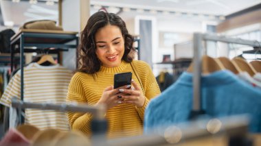 Güzel Gülümseyen Kadın Müşteri Alışverişi, Akıllı Telefon Kullanımı, İnternet Taraması, İnternette Karşılaştırma, Şık Kıyafetler Seçme. Moda Dükkanı, Renkli Markalar, Sürdürülebilir Tasarımlar
