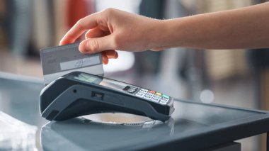 Giyim mağazası: Tezgâhta müşteri kablosuz NFC Bank Terminali üzerinden kredi kartıyla kıyafet satın alıyor. Şık Tasarımcı Markaları ile Moda Dükkanı. Yakından El 'e Odaklan.