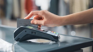 Giyim mağazası: Tezgâhta müşteri kablosuz NFC Bank Terminali üzerinden kredi kartıyla kıyafet satın alıyor. Şık Tasarımcı Markaları ile Moda Dükkanı. Yakından El 'e Odaklan.