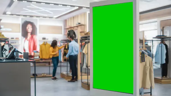 Shot of a Floor-Standing LCD Touch Screen Display with Green Screen Chroma Key Mock Up Standing in Clothing Store. Diverse People in Fashionable Shop, Choosing and Buying Stylish Clothes.
