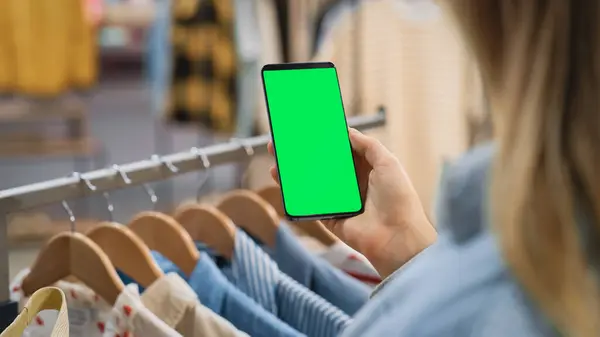 Clothing Store: Female Using Smartphone with Chroma Key Green Screen Display. Clothes Hanger with Stylish Branded Items for Retail Sale In the Background. Close Up Shot of a Mobile Device.