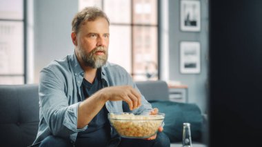 Yakışıklı, orta yaşlı sakallı adam evdeki koltukta oturup televizyon seyrediyor. Odaklanan Hayranlar Atıştırmalık Yiyor, TV Şovları İzliyor, Spor Etkinliği, Film, İstenilen Yayın Servisi.