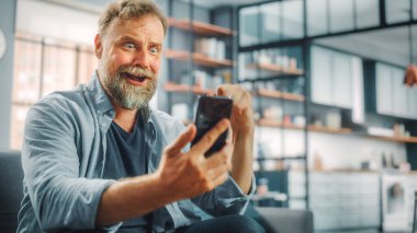 Televizyonda Kanepede Oturan Karizmatik Sakallı Adam Smartphone uygulamasını Score, Online Bet, İstatistikler, Takım Şampiyonluğu 'nu Kazandığında Zaferi Kutluyor kullanıyor. Happy Fan Sporu İzliyor