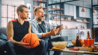 Home Two Fans on Couch Watch Basketball Maçı 'nda, Skoru Kontrol Etmek için Akıllı Telefon Uygulaması, Online Bet, Takım Şampiyonluğu' nu Kazandığında Kutla. Arkadaşlar Favori Kulüp Oyunu İzler.