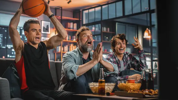 At Home Three Joyful Basketball Fans Sitting on a Couch Watch Game on TV, Celebrate Victory when Sports Team Wins Championship. Group of Friends Cheer When Favourites Play.