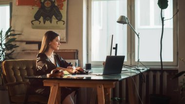 Otantik Çatı katında Yaratıcı Ajans 'ta dizüstü bilgisayarda Genç Güzel Kadın Çalışmaları. Ev Bitkileri, Sanatsal Posterleri ve Büyük Pencereli Yenilenmiş Moda Tasarımı.