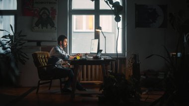 Genç Yakışıklı, çatı katındaki Yaratıcı Ajans 'ta dizüstü bilgisayarda çalışıyor. Kalemle Not Defteri Yazıyor. Ev Bitkileri, Sanatsal Posterleri ve Büyük Yuvarlak Pencereli Yenilenmiş Moda Dekorasyonu.