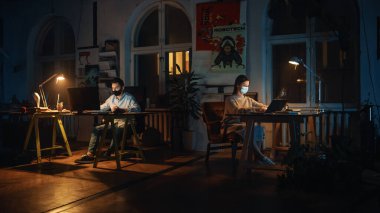 Çatı katında Yaratıcı Ajans 'ta bilgisayar üzerinde çalışırken koruyucu yüz maskesi takan iki şık çalışan. Meslektaşlar E-postalara cevap verir ve Pazarlama Projelerini yönetir. Salgın Covid-19 Konsepti.