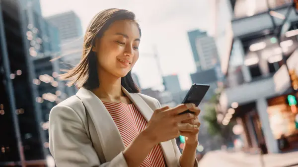 Portrait of an Attractive Japanese Female Wearing Smart Casual Clothes and Using Smartphone on the Urban Street. Manager in Big City Connecting with People Online, Messaging and Browsing Internet.