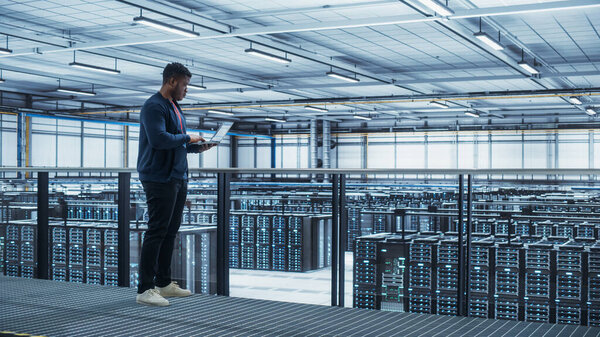 Data Center Engineer Using Laptop Computer. Server Farm Cloud Computing Specialist Facility with African American Male System Administrator Working with Data Protection Network for Cyber Security.