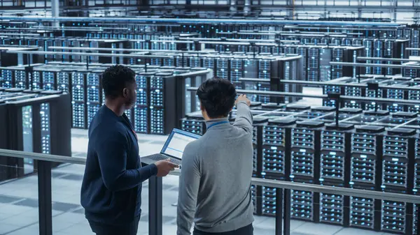 stock image Two Multiethnic Male Data Center System Administrators and IT Specialists Talk, Use Laptop. Information Technology Engineers work on Cyber Security Network Protection in Cloud Computing Server Farm.