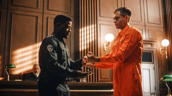 Кинематографический суд и судебное разбирательство: Грустный обвиняемый мужчина-преступник в оранжевом комбинезоне, в наручниках и с отводом охранником перед судьей и присяжными. Приговорен к тюремному заключению.