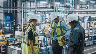 Genç Otomobil Fabrikası Mühendisleri Yüksek Görüş Yelekleri Takımı Laptop Bilgisayar kullanıyor. Otomotiv Endüstriyel Üretim Tesisi Araç Üretimi üzerine çalışıyor. Assembly Plant 'ta Çeşitlilik.