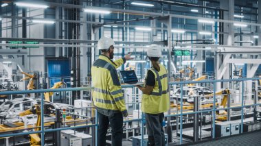 Erkek Uzman ve Kadın Araba Fabrikası Yüksek Görüş Yelekleri Mühendisi Dizüstü bilgisayar kullanıyor. Otomotiv Endüstriyel Üretim Tesisi Araç Üretimi üzerine çalışıyor. Assembly Plant 'ta Çeşitlilik.