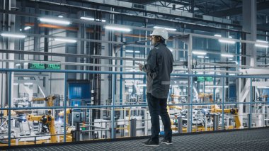 Araba Fabrikası Mühendisi Üniformalı Tablet Bilgisayar kullanıyor. Otomotiv Endüstriyel Üretim Tesisi Robotik Silah Teknolojisi ile Araç Üretimi üzerine çalışıyor. Otomatik Kurulum Tesisi.