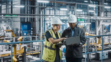 Erkek Uzman ve Kadın Araba Fabrikası Mühendisi Laptop ve Tablet Bilgisayarlar üzerinde konuşuyor ve çalışıyor. Otomotiv Endüstrisi 4.0 Tesisi Araç Üretimi üzerine çalışıyor. Assembly Plant 'ta Çeşitlilik.