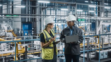 Erkek Uzman ve Kadın Araba Fabrikası Mühendisi Laptop ve Tablet Bilgisayarlar üzerinde konuşuyor ve çalışıyor. Otomotiv Endüstrisi 4.0 Tesisi Araç Üretimi üzerine çalışıyor. Assembly Plant 'ta Çeşitlilik.