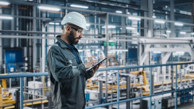 Araba Fabrikası Mühendisi Üniformalı Tablet Bilgisayar kullanıyor. Otomotiv Endüstrisi 4.0 Üretim Tesisi Robotik Silah Teknolojisi ile Araç Üretimi üzerine çalışıyor. Otomatik Kurulum Tesisi.