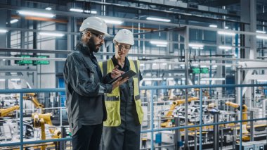 Erkek Uzman ve Kadın Araba Fabrikası Yüksek Görüş Yelekleri Mühendisi Tablet Bilgisayar kullanıyor. Otomotiv Endüstriyel Üretim Tesisi Araç Üretimi üzerine çalışıyor. Assembly Plant 'ta Çeşitlilik.