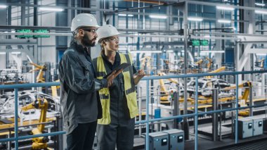 Erkek Uzman ve Kadın Araba Fabrikası Yüksek Görüş Yelekleri Mühendisi Tablet Bilgisayar kullanıyor. Otomotiv Endüstriyel Üretim Tesisi Araç Üretimi üzerine çalışıyor. Assembly Plant 'ta Çeşitlilik.