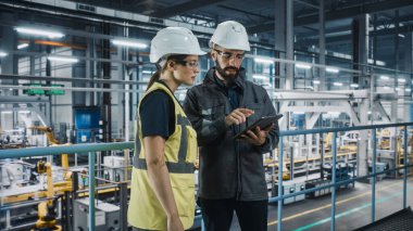 Erkek Uzman ve Kadın Araba Fabrikası Yüksek Görüş Yelekleri Mühendisi Tablet Bilgisayar kullanıyor. Otomotiv Endüstriyel Üretim Tesisi Araç Üretimi üzerine çalışıyor. Assembly Plant 'ta Çeşitlilik.