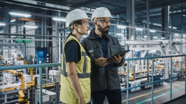 Erkek Uzman ve Kadın Araba Fabrikası Yüksek Görüş Yelekleri Mühendisi Tablet Bilgisayar kullanıyor. Otomotiv Endüstriyel Üretim Tesisi Araç Üretimi üzerine çalışıyor. Assembly Plant 'ta Çeşitlilik.