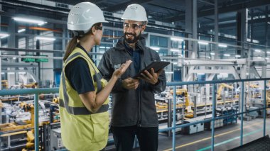 Erkek Uzman ve Kadın Araba Fabrikası Yüksek Görüş Yelekleri Mühendisi Tablet Bilgisayar kullanıyor. Otomotiv Endüstriyel Üretim Tesisi Araç Üretimi üzerine çalışıyor. Assembly Plant 'ta Çeşitlilik.