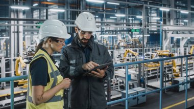 Erkek Uzman ve Kadın Araba Fabrikası Yüksek Görüş Yelekleri Mühendisi Tablet Bilgisayar kullanıyor. Otomotiv Endüstriyel Üretim Tesisi Araç Üretimi üzerine çalışıyor. Assembly Plant 'ta Çeşitlilik.