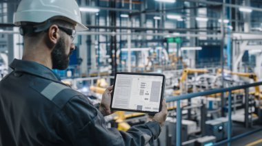 Otomobil Fabrikası Üniformalı Mühendisi Tablet Bilgisayar kullanarak Elektrik Şeması ve Şeması kullanıyor. Otomotiv Sanayi Üretim Tesisi Erkek Uzmanı Otomobil Üretimi üzerine çalışıyor.