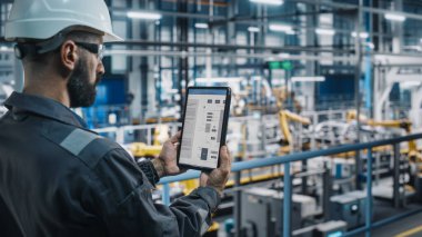 Otomobil Fabrikası Üniformalı Mühendisi Tablet Bilgisayar kullanarak Elektrik Şeması ve Şeması kullanıyor. Otomotiv Sanayi Üretim Tesisi Erkek Uzmanı Otomobil Üretimi üzerine çalışıyor.