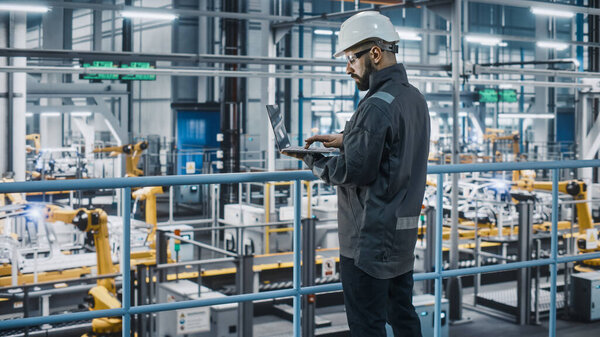 Car Factory Engineer in Work Uniform Using Laptop Computer. Automotive Industrial Manufacturing Facility Working on Vehicle Production with Robotic Arms Technology. Automated Assembly Plant.