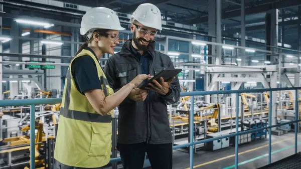 Erkek Uzman ve Kadın Araba Fabrikası Yüksek Görüş Yelekleri Mühendisi Tablet Bilgisayar kullanıyor. Otomotiv Endüstriyel Üretim Tesisi Araç Üretimi üzerine çalışıyor. Assembly Plant 'ta Çeşitlilik.