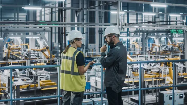 Erkek Uzman ve Kadın Araba Fabrikası Yüksek Görüş Yelekleri Mühendisi Dizüstü bilgisayar kullanıyor. Otomotiv Endüstriyel Üretim Tesisi Araç Üretimi üzerine çalışıyor. Assembly Plant 'ta Çeşitlilik.