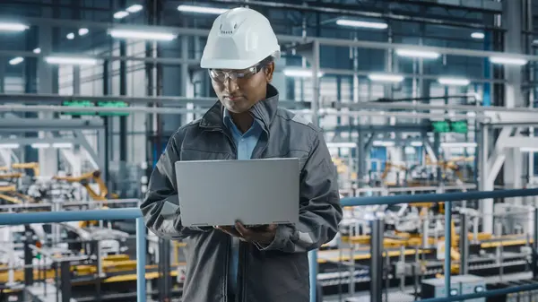 Portrait of Automotive Industry Engineer in Safety Uniform Using Laptop at Car Factory Facility. Happy Assembly Plant Multiethnic Man Specialist Working on Manufacturing Modern Electric Vehicles.