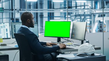 Araba Fabrikası: Siyah Mühendis Bilgisayarda Çalışıyor, İki Monitör Kroma Anahtar Yeşil Ekran ve Otomatik Robot Silahlanma Hattı Üretim Hattı Yüksek Teknolojik Elektrikli Araçlar